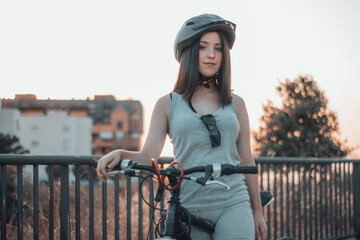 Muchacha joven parada y aparcada con una bicicleta y casco en la ciudad junto a los arboles y la naturaleza de un parque - obrazy, fototapety, plakaty