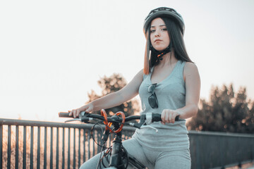 Muchacha joven parada y aparcada con una bicicleta y casco en la ciudad junto a los arboles y la...