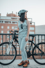 Joven adolescente parada en la ciudad junto con una bicicleta y casco haciendo ejercicio - obrazy, fototapety, plakaty