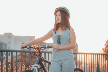 Joven adolescente parada en la ciudad junto con una bicicleta y casco haciendo deporte - obrazy, fototapety, plakaty