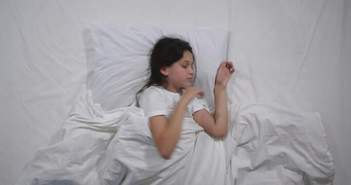  Top view  of girl 9 years old with long brown sleeping  in bed with white linens in light room. High angle shooting. Teenager girl having restless sleep. Good morning 4k 