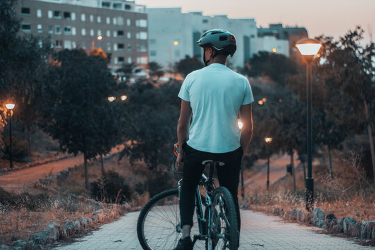 Hombre joven paseando con una bicicleta de espaldas con un casco de protecci&oacute;n en un parque al anochecer