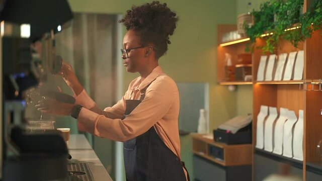 Barista African-American woman at coffeemachine. Spbas Black lady worker cleans contemporary equipment with hot steam and napkin on counter in coffee shop