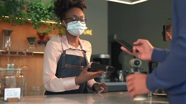 Takeaway coffee safe serving. Spbas Young Afro-American lady bartender in medical mask gives beverage in to-go cup and terminal to man to pay in coffee shop