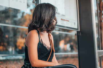 Chica adulta de pelo castaño esperando en la parada del autobús sentada para realizar el transporte publico - obrazy, fototapety, plakaty