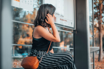 Chica adulta de pelo castaño esperando en la parada del autobús sentada para realizar el transporte publico - obrazy, fototapety, plakaty