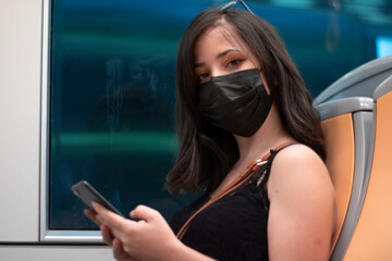 Chica joven de pelo oscuro sentada en el interior metro con la mascarilla y observando el teléfono...