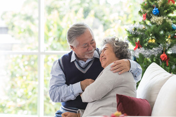 Happy Asian senior man embracing his wife on Christmas day at home. Senior couple spending time together with Christmas tree. Merry Christmas and Happy Holidays. Christmas holiday celebration