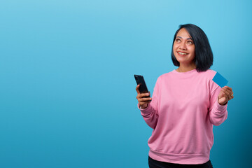 Portrait of beautiful smiling asian woman using mobile phone and holding credit card and looking up empty space