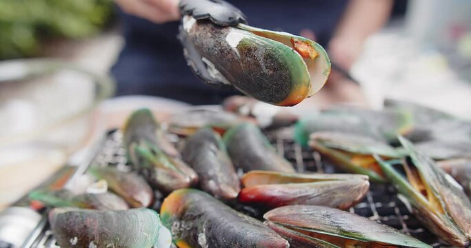 person roasting fresh juicy Green clam Shell mussels with tongs grilling on a hot gas stove, cooking seafood party outdoor