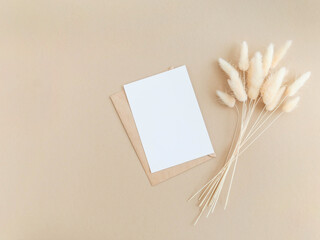 White blank card for text and envelope next to bouquet of dry Lagurus on beige background.