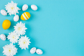 White flowers and painted Easter eggs on a blue background. Festive floral concept with clean text space. Flat lay. View from above.