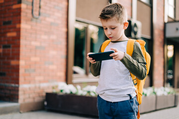 Back to school. Cute child with backpack and mobile phone going to school. Boy pupil with bag. Elementary school student going to classes. Kid walking outdoors on the city street after class.