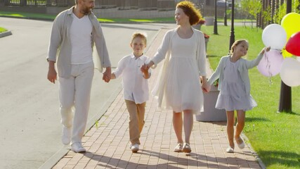 Happy caucasian family walking together along the street on summer day. Joyous parents smiling and talking to kids, little girl holding bunch of colorful balloons