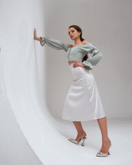 Elegant woman wearing pastel green top, silver silk skirt and high heels standing at white gray background and leaning on wall. Pretty fashion female model with pony tail brunette hair