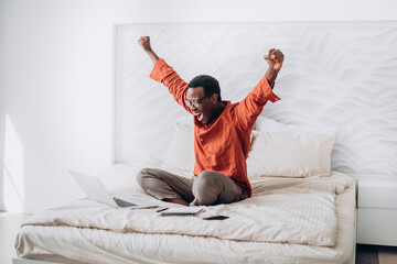 Joyful African-American guy in casual clothes and glasses puts hands up sitting near open laptop on comfortable bed at home