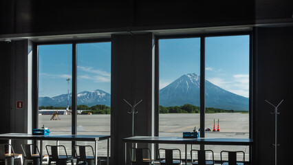 Conical volcanoes against the blue sky are visible through the airport windows. There is snow on the slopes.  In the foreground are tables and chairs of the cafe. Petropavlovsk Kamchatsky