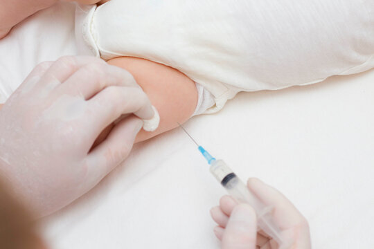 The doctor's hands in rubber protective gloves hold a syringe. An infant receiving the vaccine in the leg. Visit to the pediatrician in the hospital. A two-month-old baby. To close.