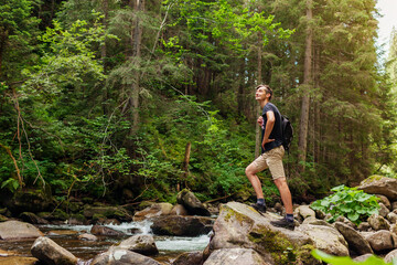 Naklejka na ściany i meble Traveler hiker with backpack enjoys landscape by mountain river in Carpathian forest. Trip to summer Ukraine