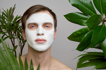 Portrait of young handsome man with white cosmetic mask on his face against background of green...