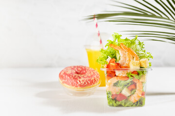 Wellness fresh summer food take away - set of orange juice, pink donut, tropical salad with shrimps, lettuce, red pepper, croutons in plastic box in white bar interior with marble tile, palm leaf.