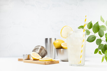 Healthy vitamin lemon drink cooking in soft light white kitchen with marble tile, green leaves in sunlight or outdoor  - fresh natural lemonade with ice, ingredients on white wood table.