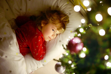 Little cute toddler girl in bed under Christmas tree and dreaming of Santa at home, indoors. Traditional Christian festival. Happy kid child waiting for gifts on xmas. Cozy soft light