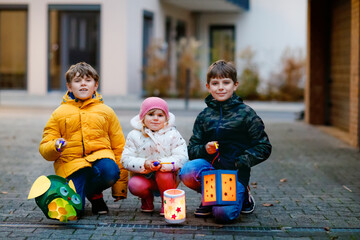 Little toddler girl and two kids boys holding selfmade lanterns with candle for St. Martin procession. Three Healthy children happy about family parade in kindergarten. German tradition Martinsumzug