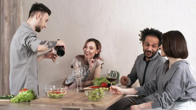 Young man pouring red wine into glasses for friends while they smiling and chatting on dinner party at home