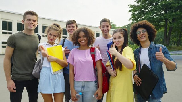Group of diverse students in casual wear with backpacks on shoulders and books in hands looking at camera and showing thumbs up, college on background. Cheerful friends happy together