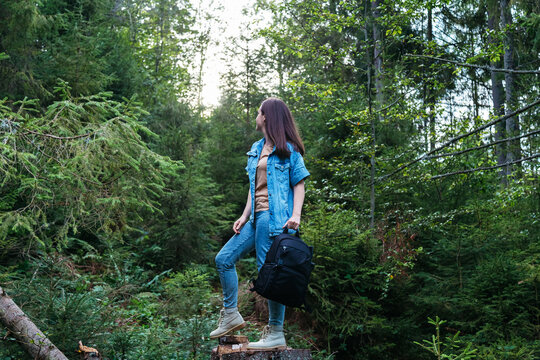 Woman in denim suit in boots with Photo backpack is engaged in hiking in forest. Unrecognizable caucasian young woman walking in nature in park. Journey alone back view