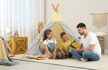 Father reading book to children near toy wigwam at home © New Africa