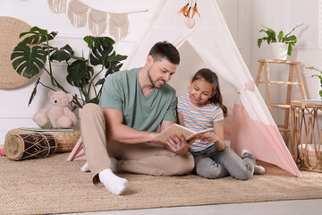 Father and daughter reading book near toy wigwam at home © New Africa