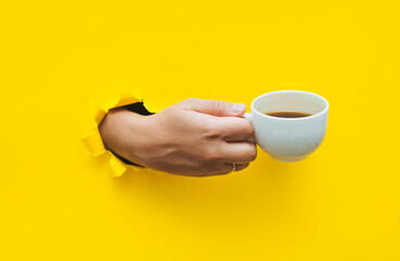 A right man's hand holds a white cup with coffee or tea. Torn hole in yellow paper, copy space. The...