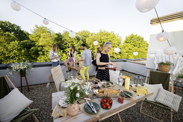 Young group of stylish people having a festive dinner on the beautifully decorated roof terrace. Friends hanging out together at picnic. Wide view