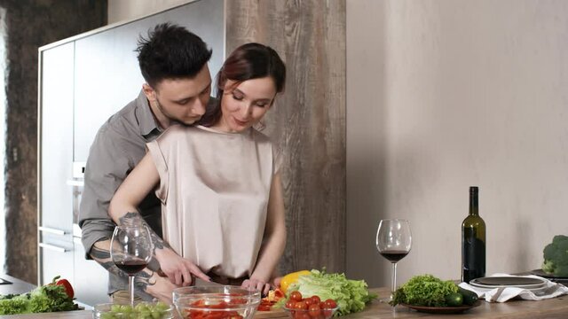 Young tattooed woman smiling and cutting vegetable for salad on kitchen table while her loving husband kissing and hugging her with affection