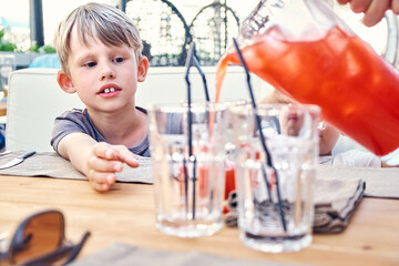 Cute junior schoolboy reaches hand for glass filled with delicious strawberry juice on wooden table in hotel cafe bar close view