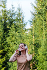 Young woman enjoys listening to music with headphones in forest in nature. Portrait young beautiful caucasian woman dreaming listening to music via headphones with closed eyes outdoor