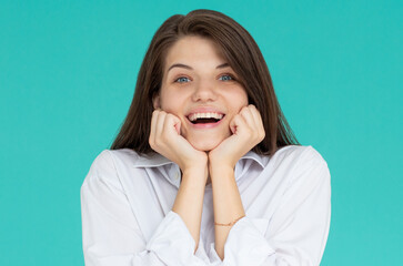 Indoor portrait of young attractive European girl isolated on blue background looking straight at camera propping up her chin with fists feeling great joy, smiling and laughing at joke