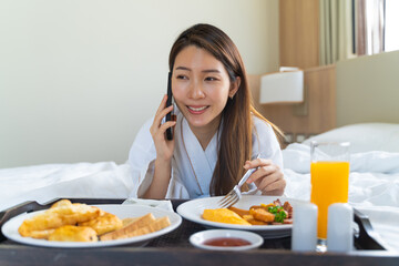 Obraz na płótnie Canvas Portrait beautiful young Asian woman wearing bathrobe talking on a phone while eating breakfast on a bed in bedroom