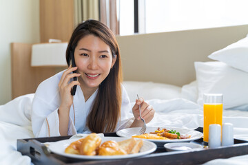 Obraz na płótnie Canvas Portrait beautiful young Asian woman wearing bathrobe talking on a phone while eating breakfast on a bed in bedroom