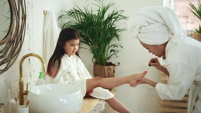 Mother daughter play beauty salon. Spbd Concentrated Asian lady applies nailpolish on toe nails of little kid sitting on counter in light spacious bathroom