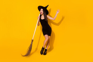 Full body photo of sing witch young lady with broom wear black dress cap shoes isolated on yellow color background