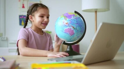 Home education of junior schoolchildren. Spbd Curious brunette schoolgirl turns globe learning geography at video lesson via laptop at table in room