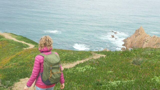 happy girl with blonde ponytail in pink jacket with large backpack walks along steep coast near azure ocean on nasty day