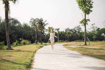 Beautiful caucasian woman with long blond hair walking barefoot in the park with palm trees. Wearing white dress. Romantic summer picture