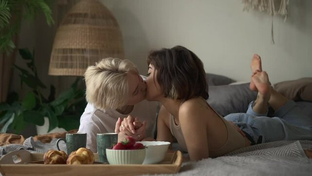 Young lesbian women kissing and smiling while lying on bed in home bedroom spbd. Closeup view of beautiful homosexual females kiss and pose with smile, show love and lie in light room, tray of food