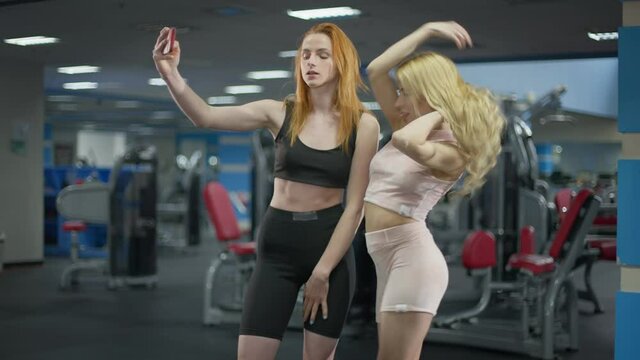 Young slim Caucasian millennial women taking selfie in gym smiling and posing. Portrait of cheerful beautiful redhead and blond millennials photographing for social media. Sport and internet addiction