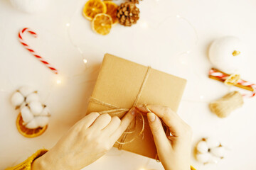 Packaging of Christmas gift box. Fir cones, dry orange slices and other decorations on table. Girl in sweater is preparing surprise for New Year. Festive atmosphere. Women's hands tie ribbon in knot.