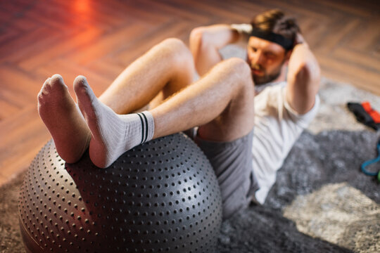 Blur view of handsome man lying on floor and doing crunches exercises for abdominal muscles. Caucasian bearded man using fitball for training at home. Focus on man's legs on fitball